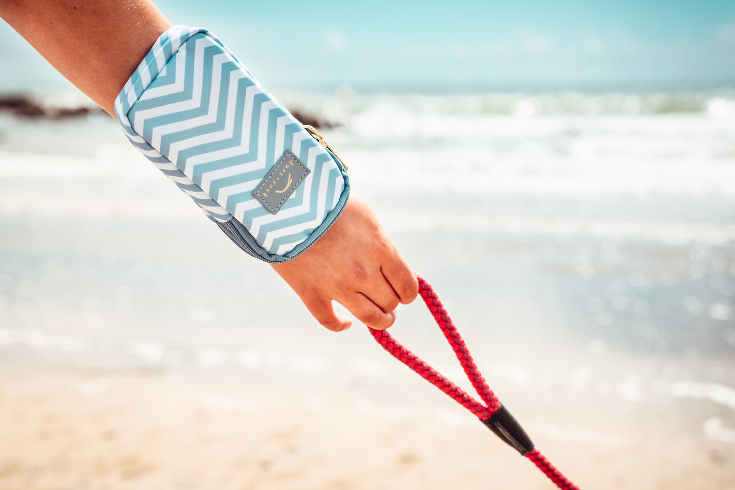 Person walking a dog on the beach while wearing the Dust and Rock Mini Wrist Pocket – 'Sea You There' edition; a compact, hands-free wrist pouch for smaller phones and essentials, perfect for seaside strolls and dog walks.