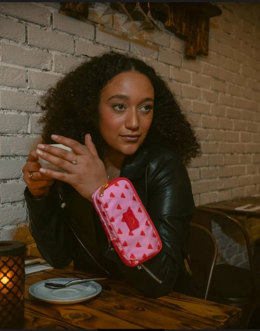 Woman wearing the Dust and Rock Original Wrist Pocket – 'Toast My Marshmallows!' edition in pink with red triangle print while enjoying coffee; stylish hands-free purse perfect for cafés, city strolls, festivals, and everyday adventures.